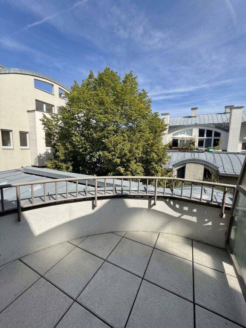 Eine offene Terrasse mit Fliesenboden und Geländer, umgeben von Bäumen und Gebäuden unter blauem Himmel.