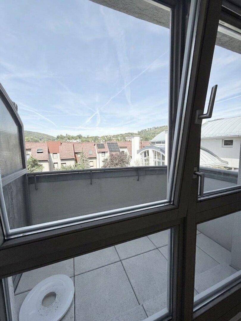 Blick aus einem Fenster auf eine geflieste Terrasse mit umliegenden Gebäuden und Hügeln im Hintergrund.