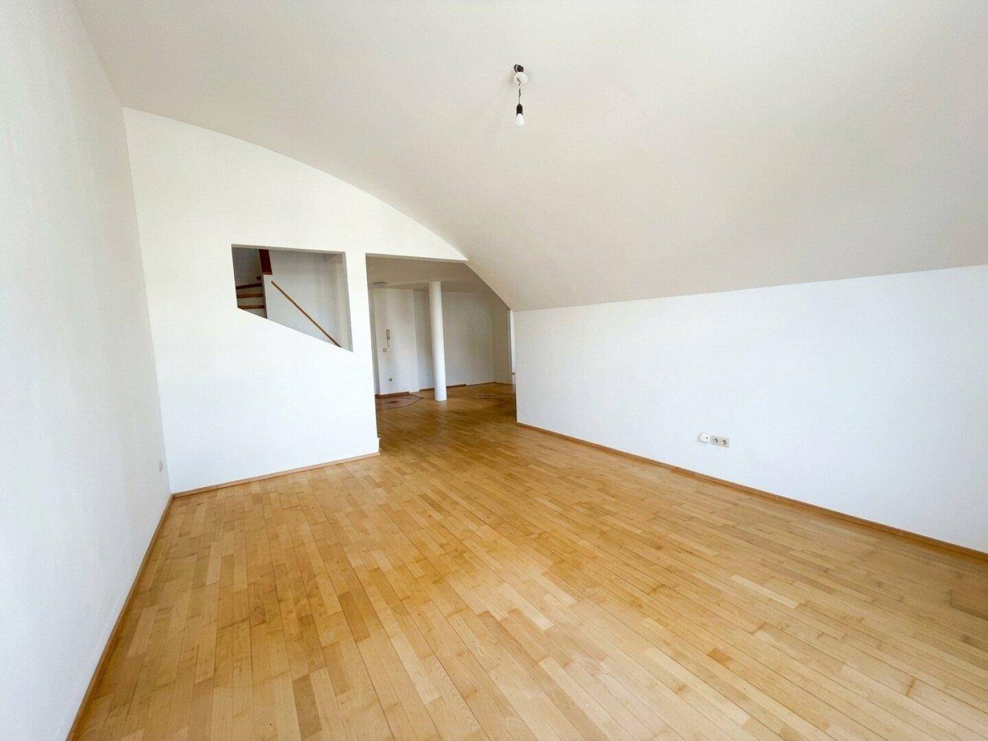 Leerer Raum mit Dachschräge und hellem Holzparkett, der auf einen weiteren Bereich führt.