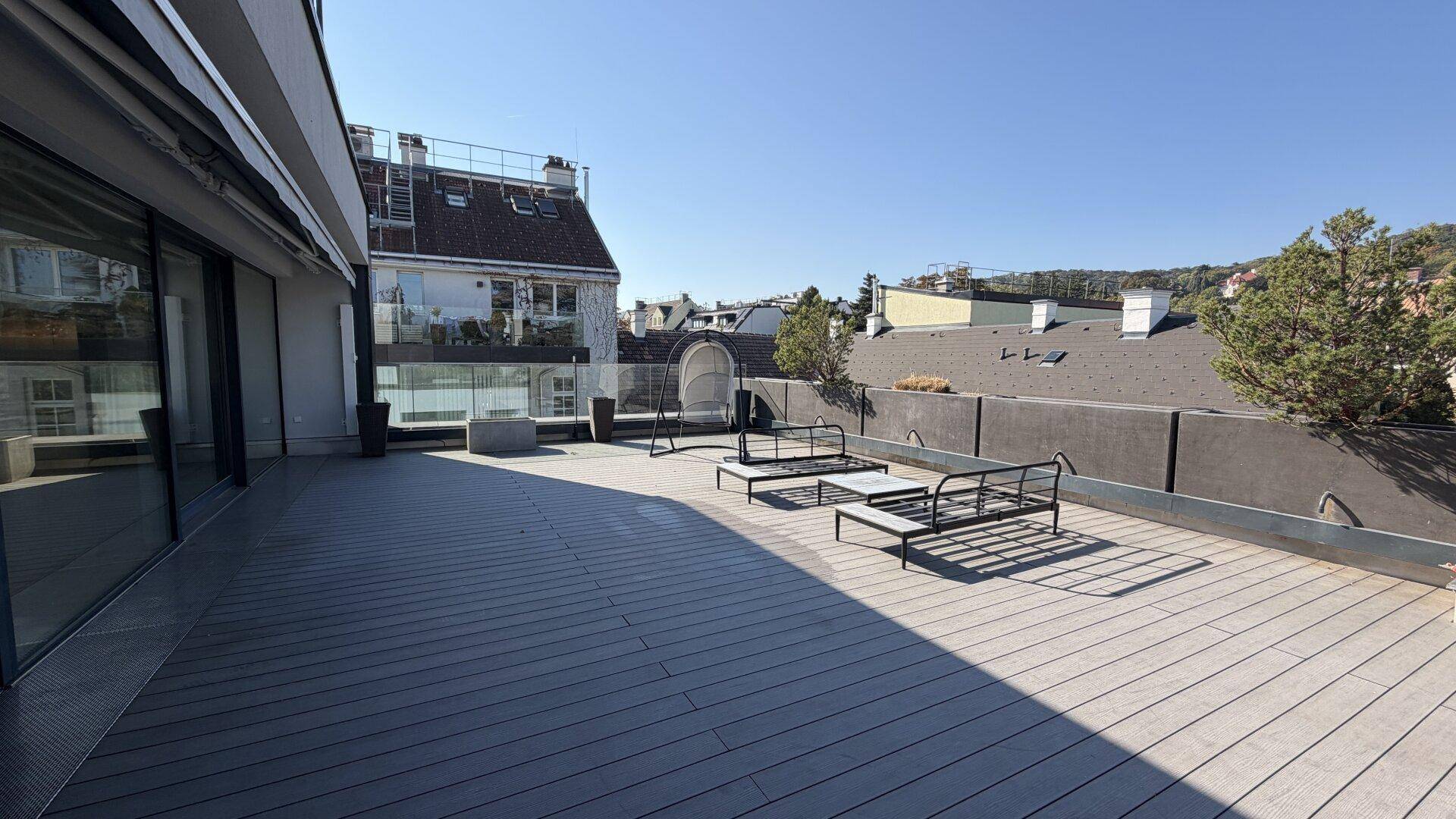Weitläufige Sonnenterrasse mit Holzdeck, Glasgeländer und Blick auf die Stadt und Bäume.