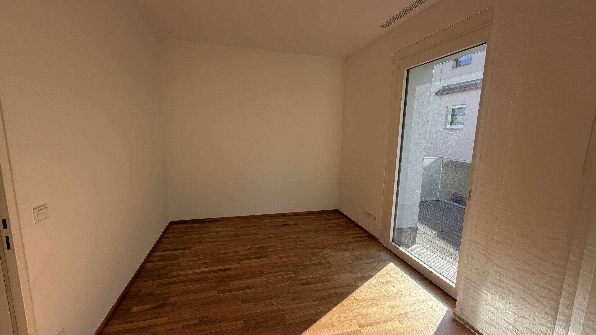 Helles Zimmer mit Parkettboden, weißen Wänden und großem Zugang zu einem Außenbereich.