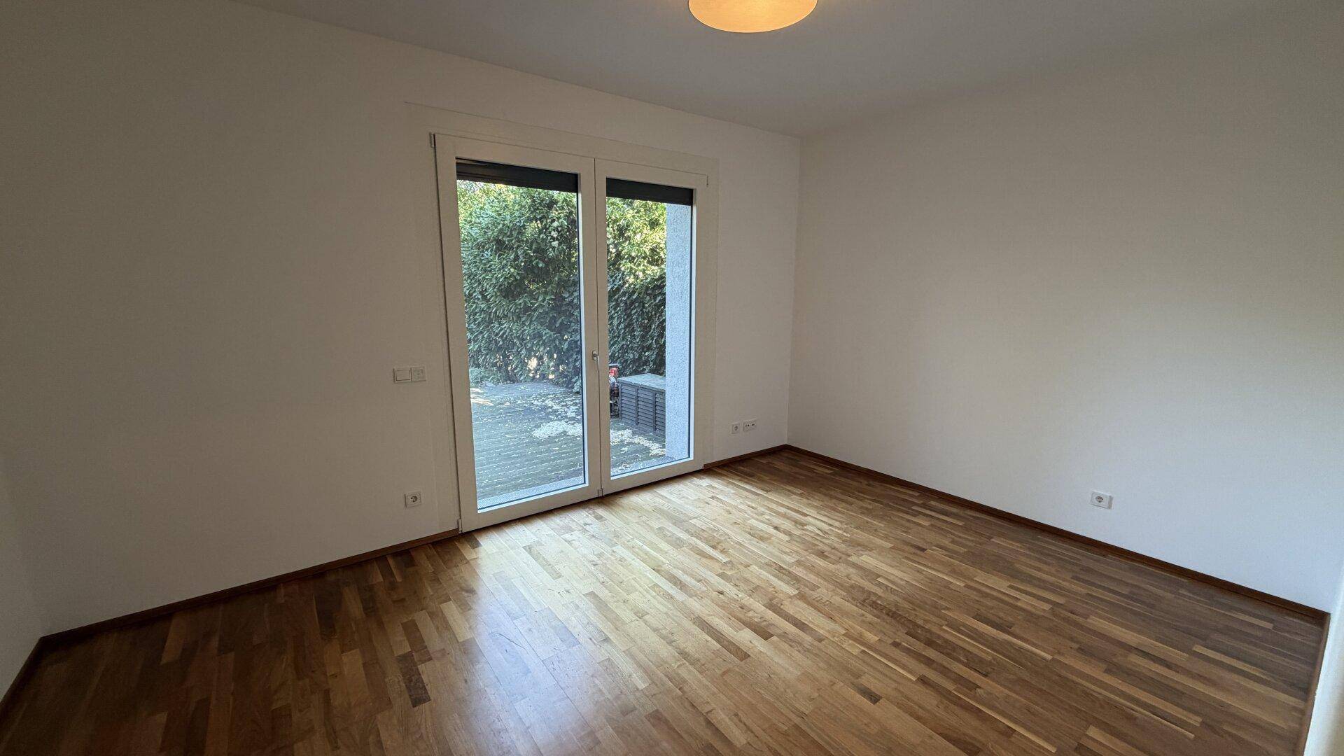 Leeres Zimmer mit Parkettboden, weißen Wänden und großem Zugang zu einem Außenbereich.