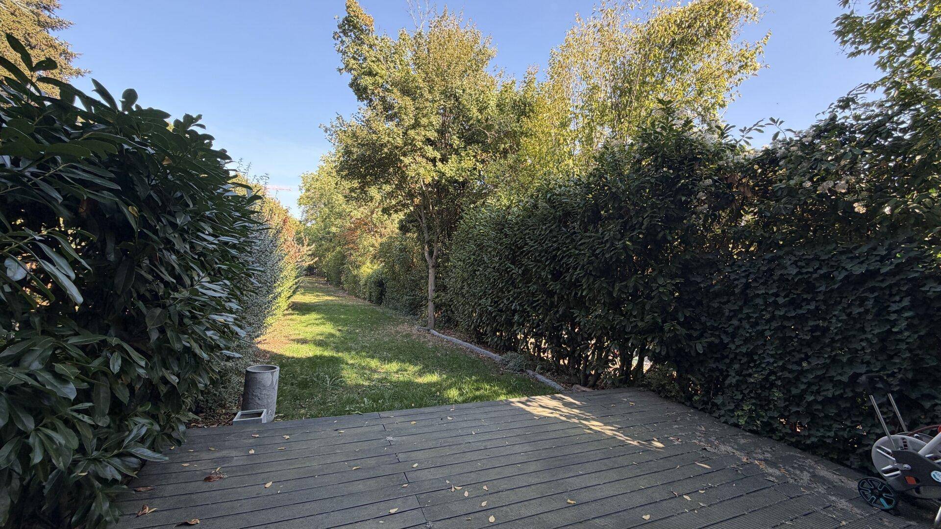 Gartenbereich mit Holzterrasse, Rasenfläche und dichten Hecken und Bäumen.