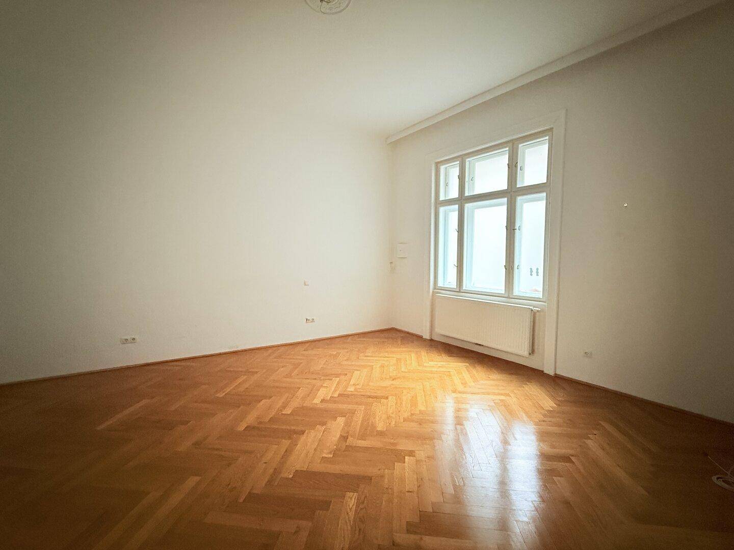 Leeres Zimmer mit Fischgrätparkett, hohen Decken und großem Fenster.