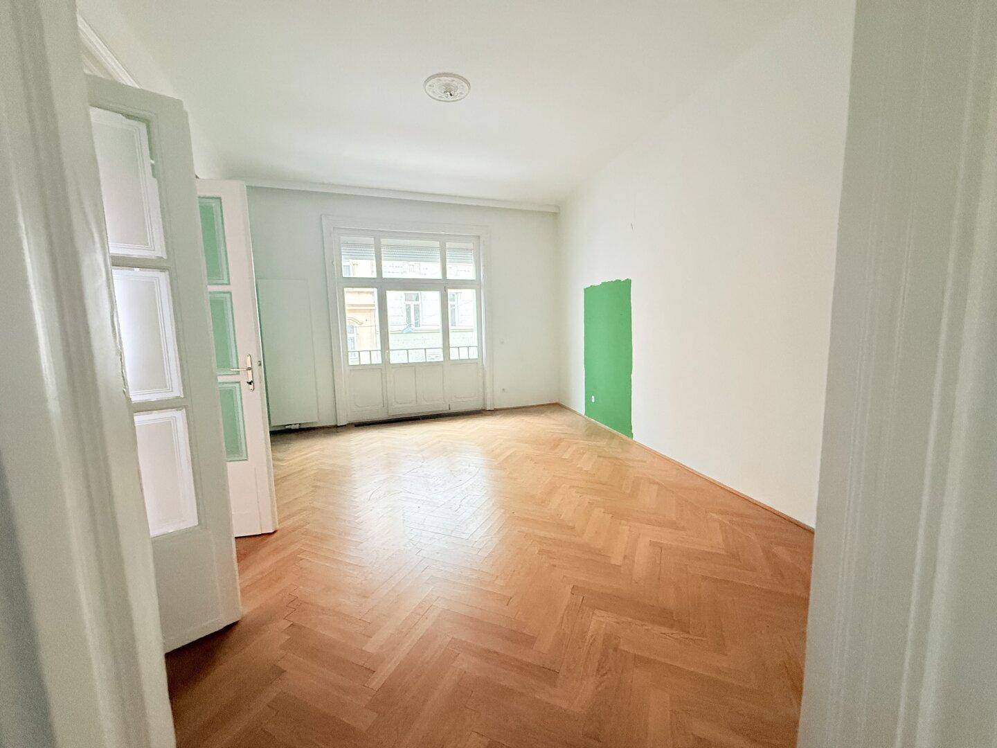 Helles Zimmer mit Fischgrätparkett, hohen Decken und einem grünen Akzent an der Wand.
