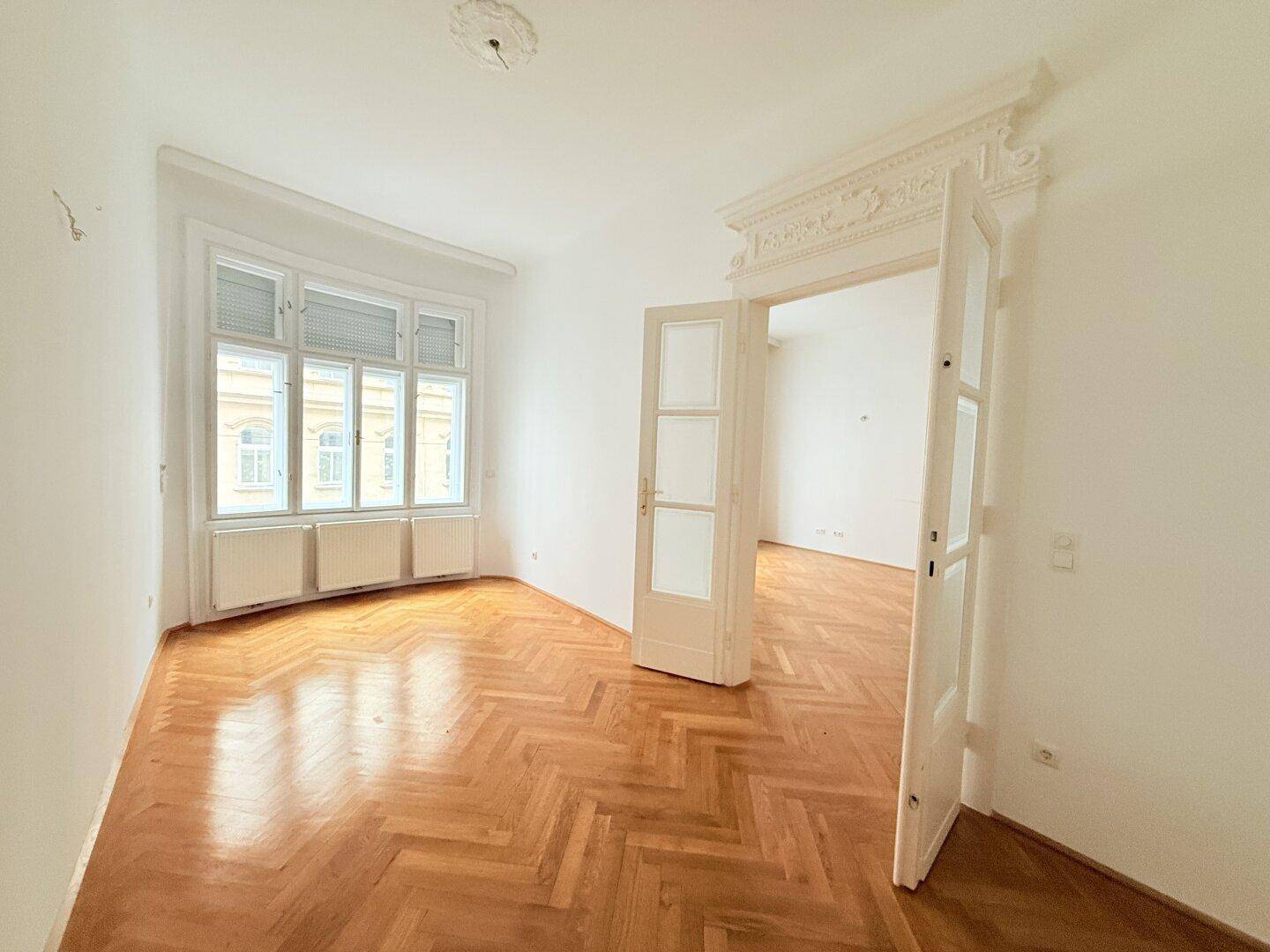 Helles Zimmer mit Fischgrätparkett, hohen Decken und großen Fenstern mit Blick nach draußen.