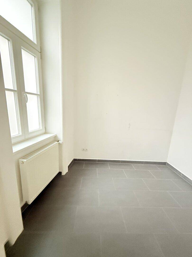 Leeres Zimmer mit Fliesenboden, Heizkörper und großem Fenster.