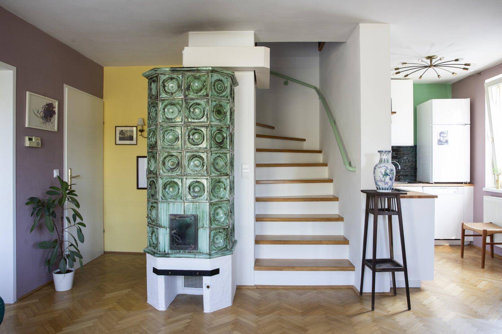 Offener Wohnbereich mit Kachelofen, Treppe zum Obergeschoss und angrenzender Küche.