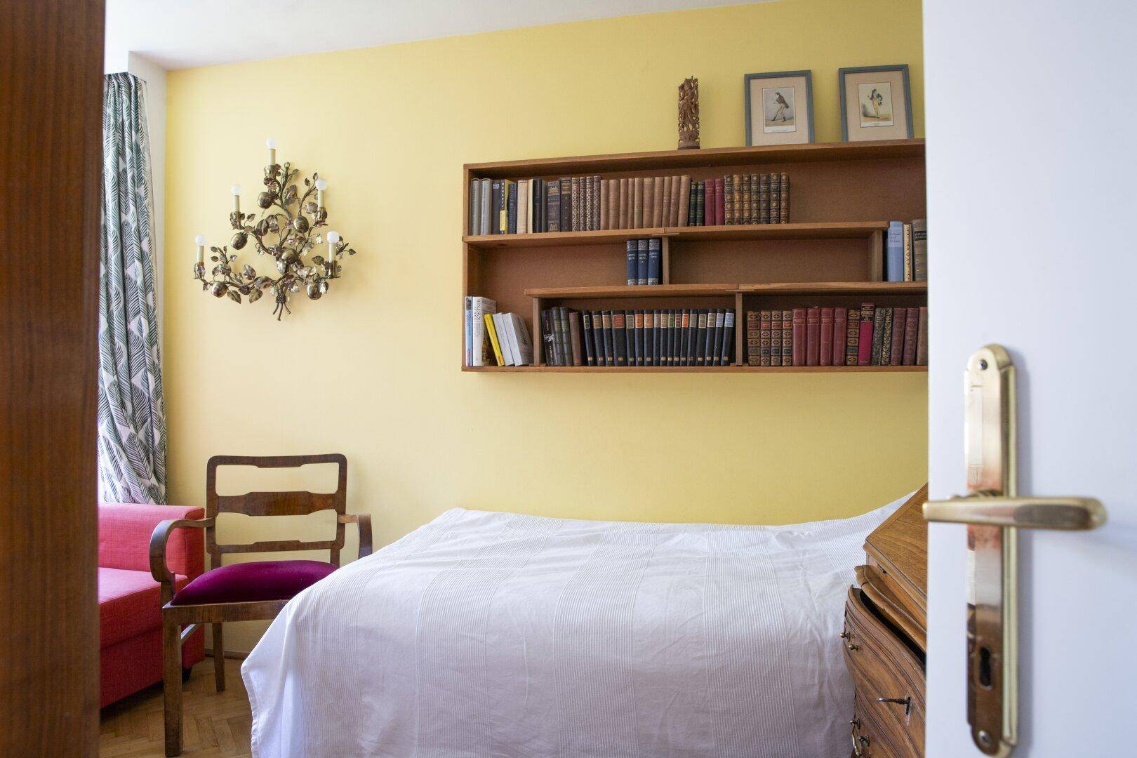 Helles Schlafzimmer mit Bett, Bücherregal und einem Sessel, durch eine offene Tür sichtbar.