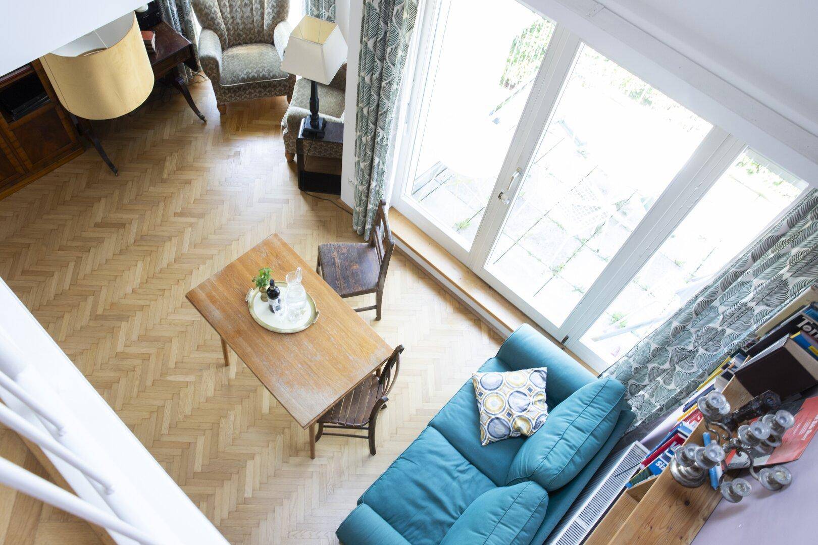 Blick von oben auf einen hellen Wohnbereich mit Esstisch, Sofa und großen Fenstern.
