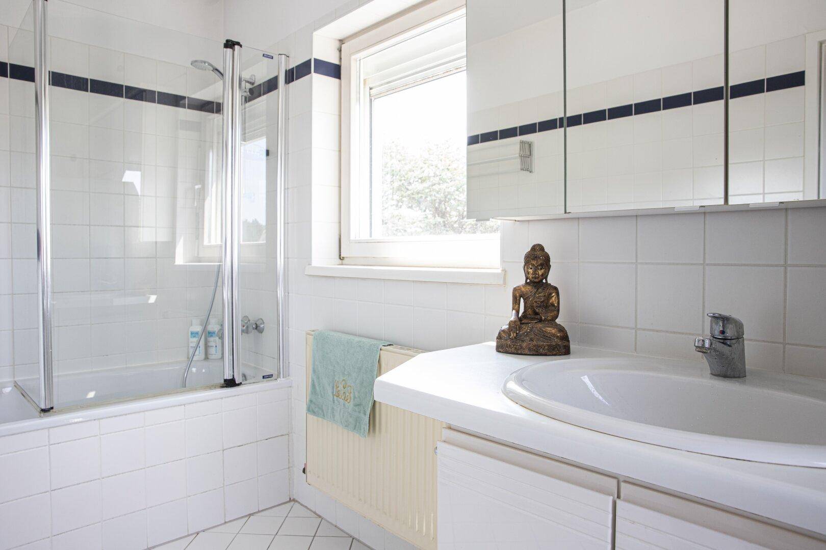 Helles Badezimmer mit Badewanne, Duschabtrennung, Waschbecken und Fenster für natürliche Belüftung.