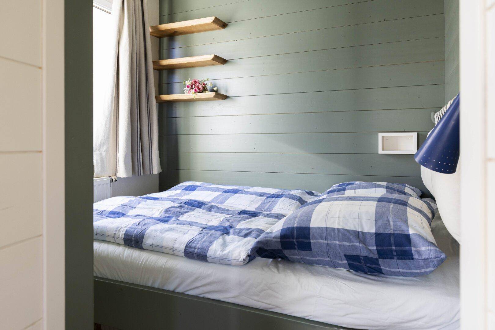 Kleiner Schlafbereich mit Einzelbett, blauer karierter Bettwäsche und Holzregalen an der Wand.