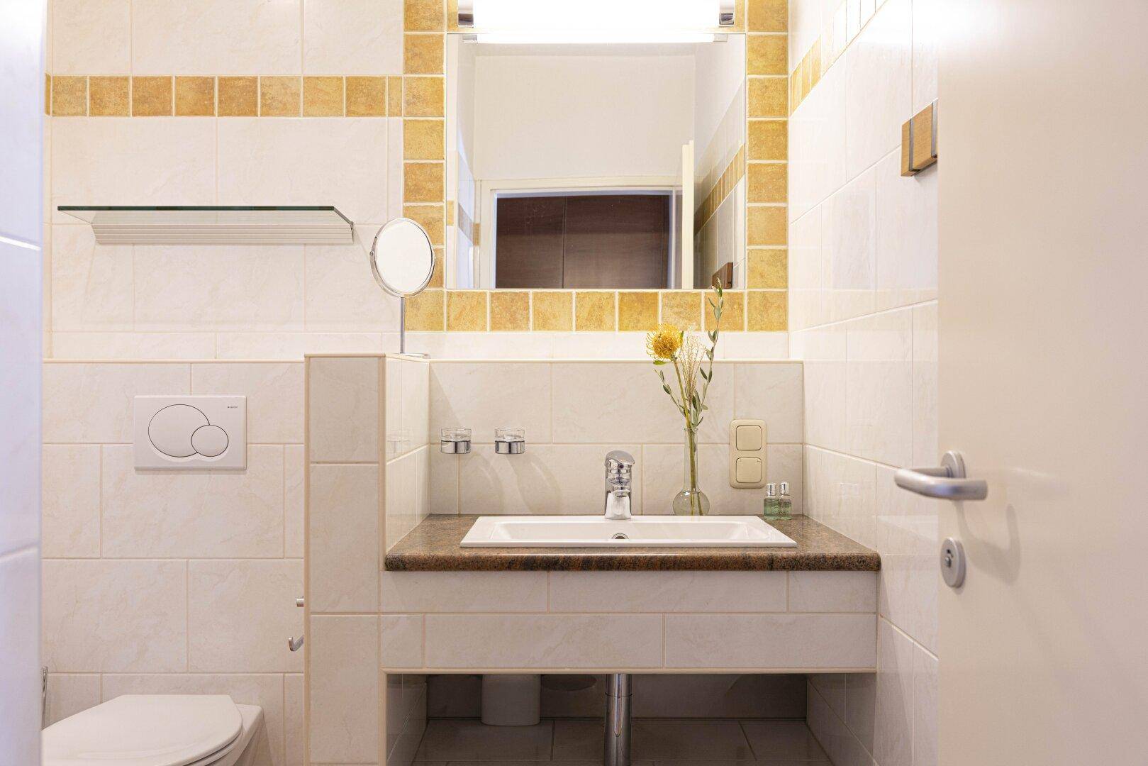 Helles Badezimmer mit Waschbecken, Spiegel und beigen Fliesen, inklusive Toilette.