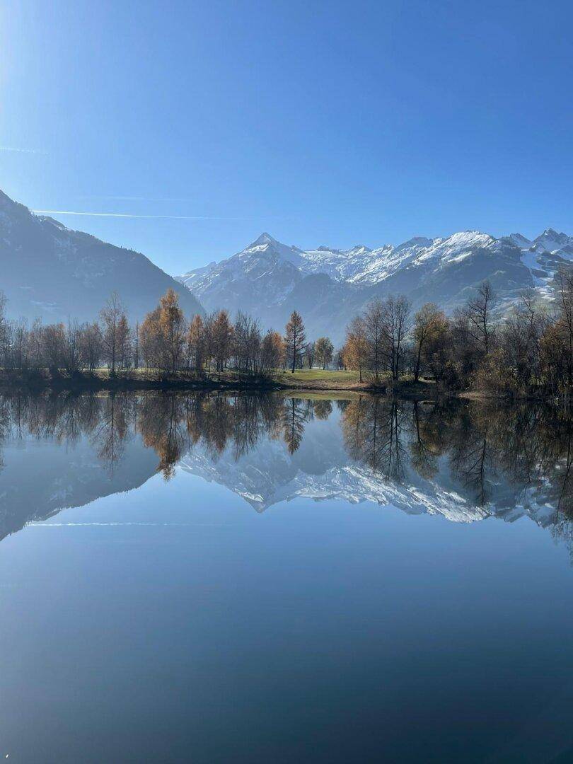 Ruhiger See mit spiegelnden Bergen und Herbstbäumen unter blauem Himmel.