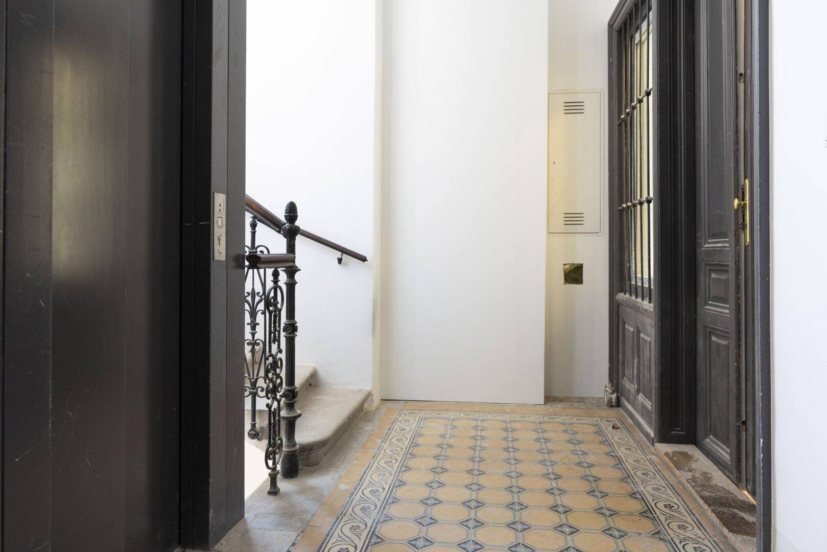 Historischer Eingangsbereich mit Aufzug, Treppe und gemustertem Fliesenboden.
