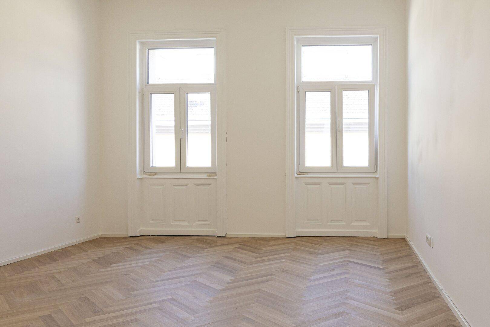 Geräumiges Zimmer mit Fischgrätparkett und hohen Fenstern, bereit zur Einrichtung.