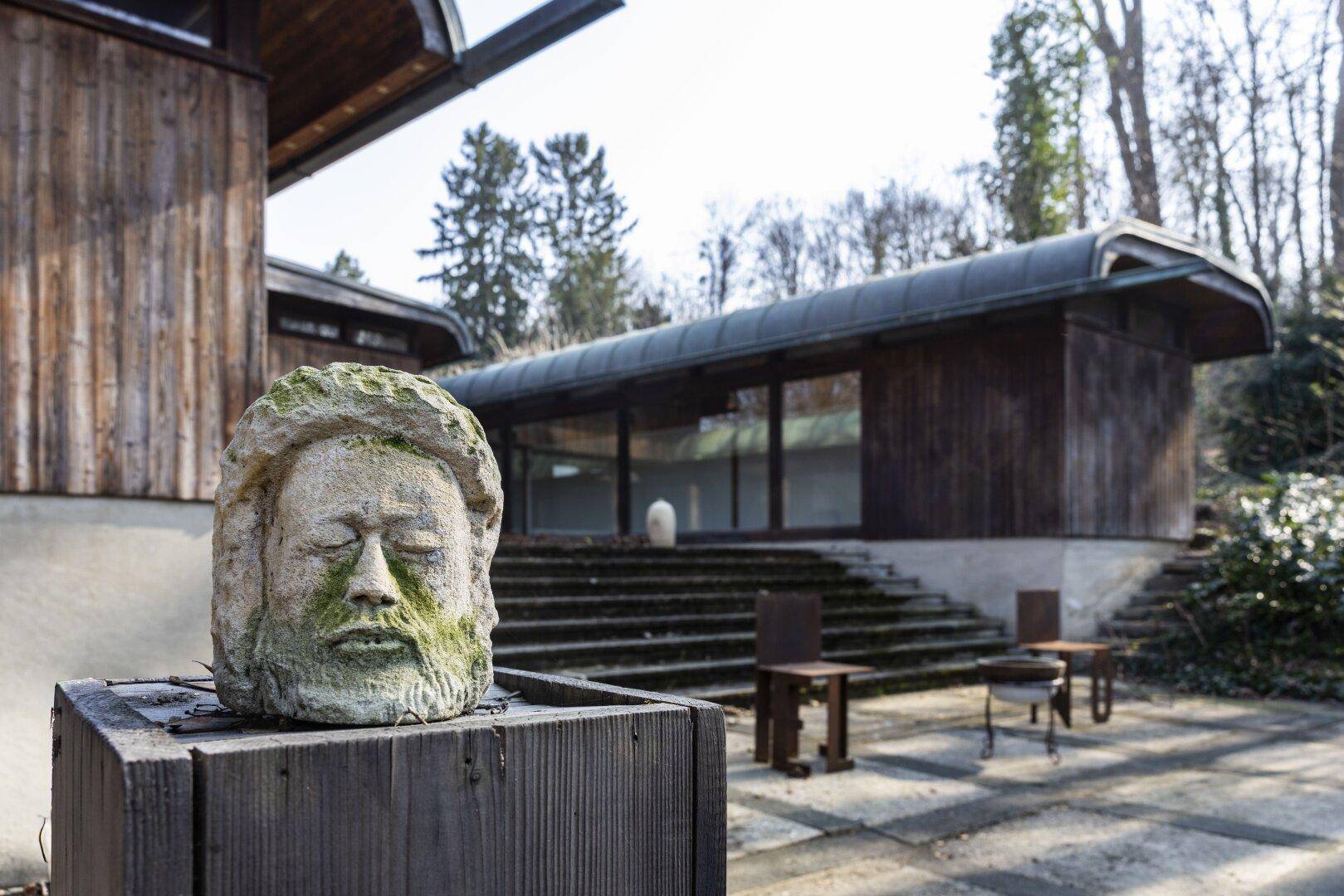 Skulptur vor der modernen Architektur mit Holzfassade und geschwungenem Dach, umgeben von Bäumen.