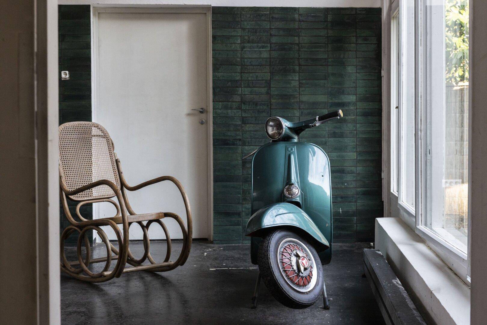 Wohnbereich mit Rattanschaukelstuhl und einer Vintage-Vespa vor der dunkelgrünen Fliesenwand.