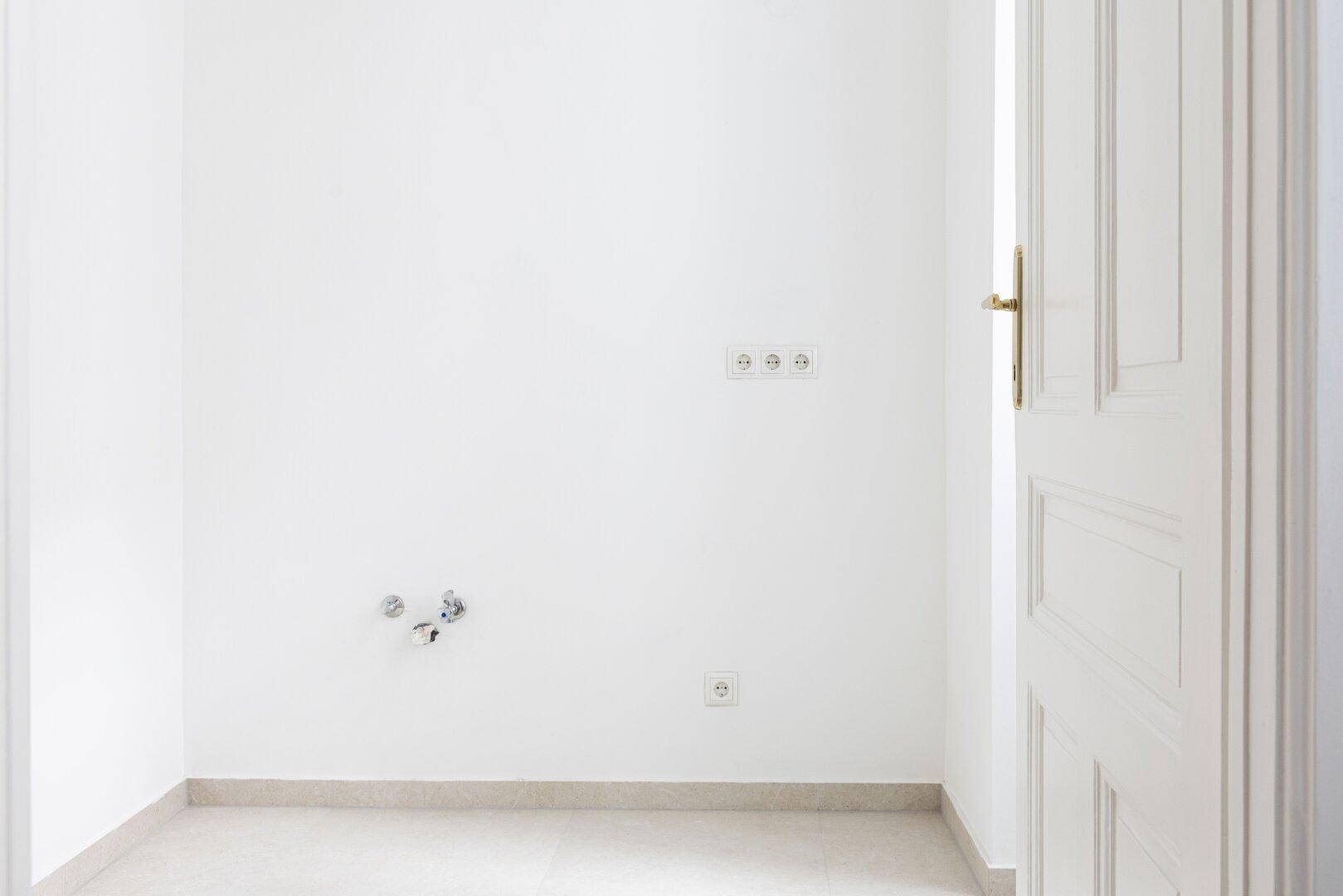 Detailansicht eines hellen Raumes mit Fliesenboden, Sanitäranschlüssen und Steckdosen an der weißen Wand.