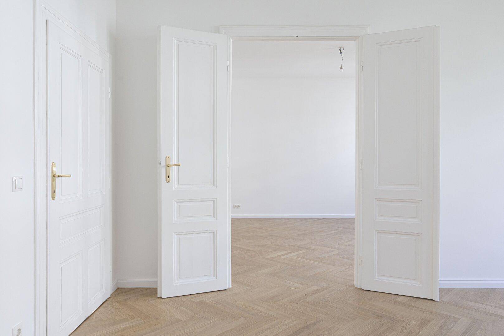 Heller Raum mit Fischgrätparkett und hohen weißen Doppeltüren, die zu einem weiteren Zimmer führen.