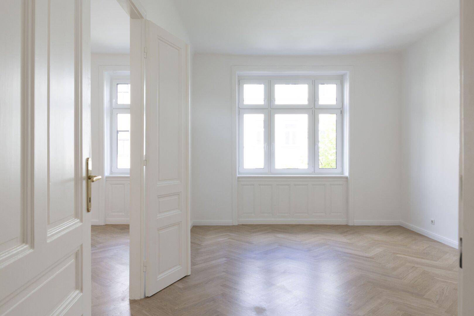 Geräumiges, lichtdurchflutetes Zimmer mit Fischgrätparkett und einem großen Fenster, das viel Tageslicht hereinlässt.