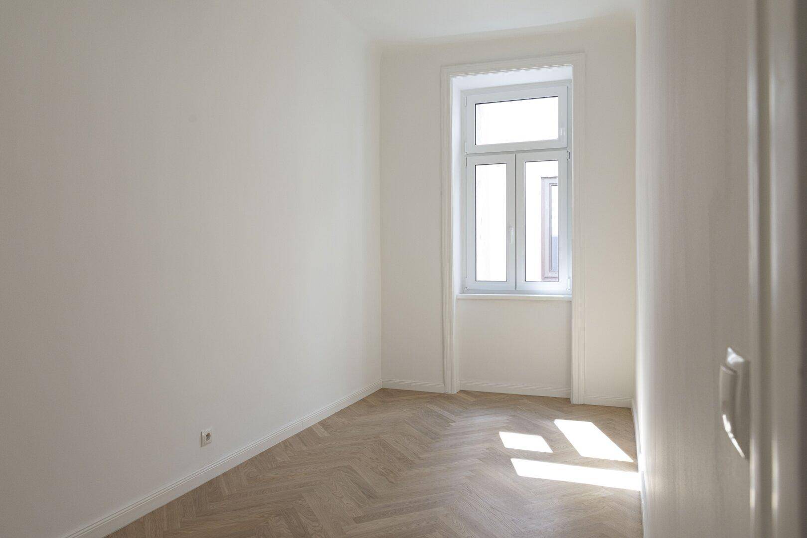 Helles Schlafzimmer mit Fischgrätparkett und einem Fenster, durch das die Sonne auf den Boden fällt.