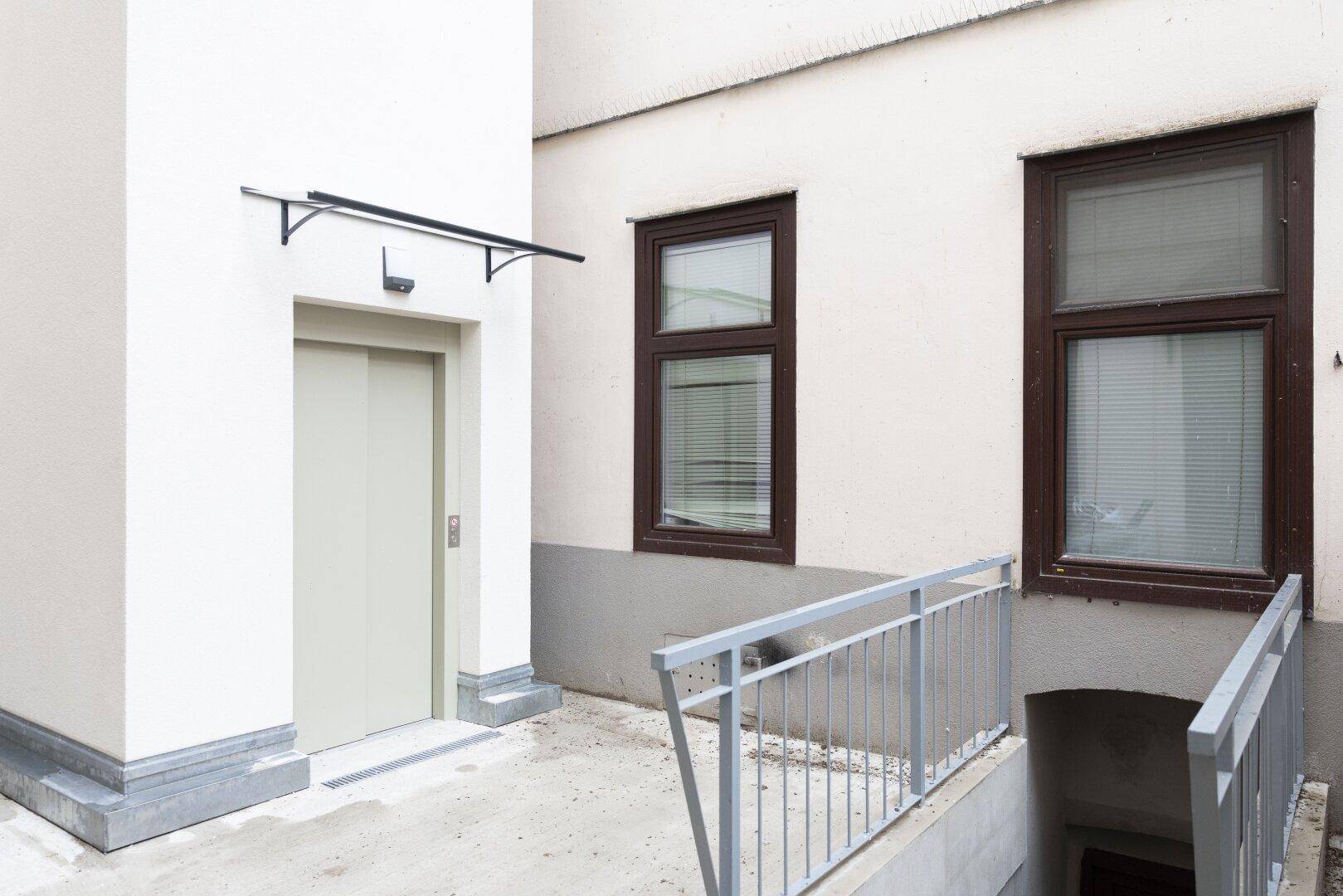 Gebäudeeingang mit modernem Aufzug, Rampe und Treppenabgang zum Kellerbereich, barrierefrei gestaltet.
