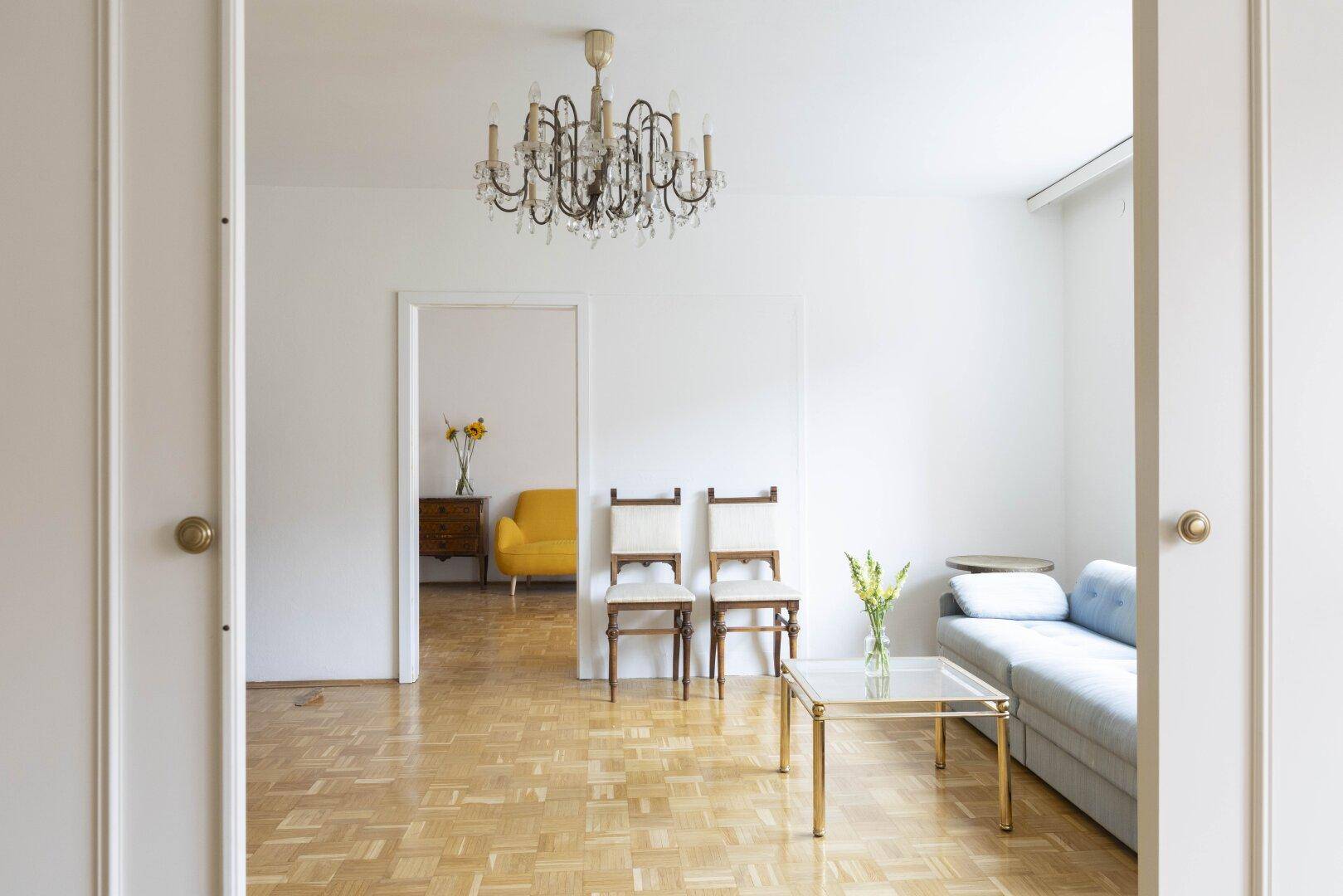 Helles Wohnzimmer mit Parkettboden, Kronleuchter und Schiebetüren zu einem weiteren Raum.