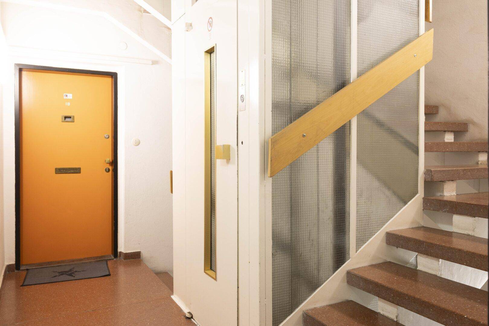 Eingangsbereich mit Aufzug, Treppe und einer orangefarbenen Wohnungstür.
