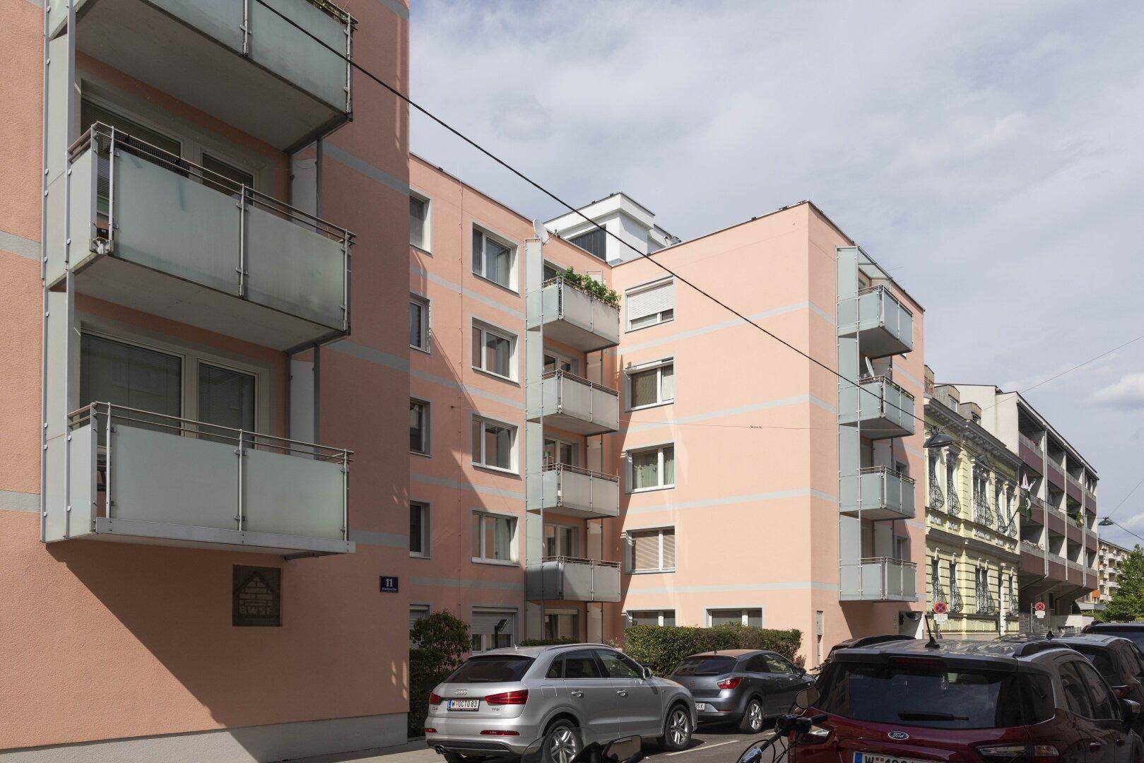 Mehrfamilienhaus mit Balkonen und parkenden Autos auf der Straße.