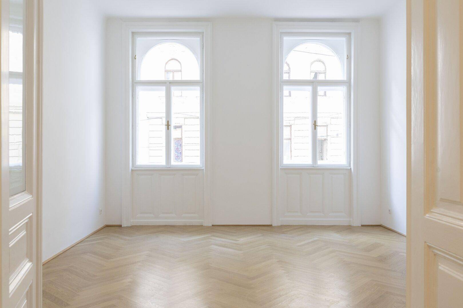 Geräumiger, heller Raum mit Fischgrätparkett und zwei großen, eleganten Bogenfenstern.