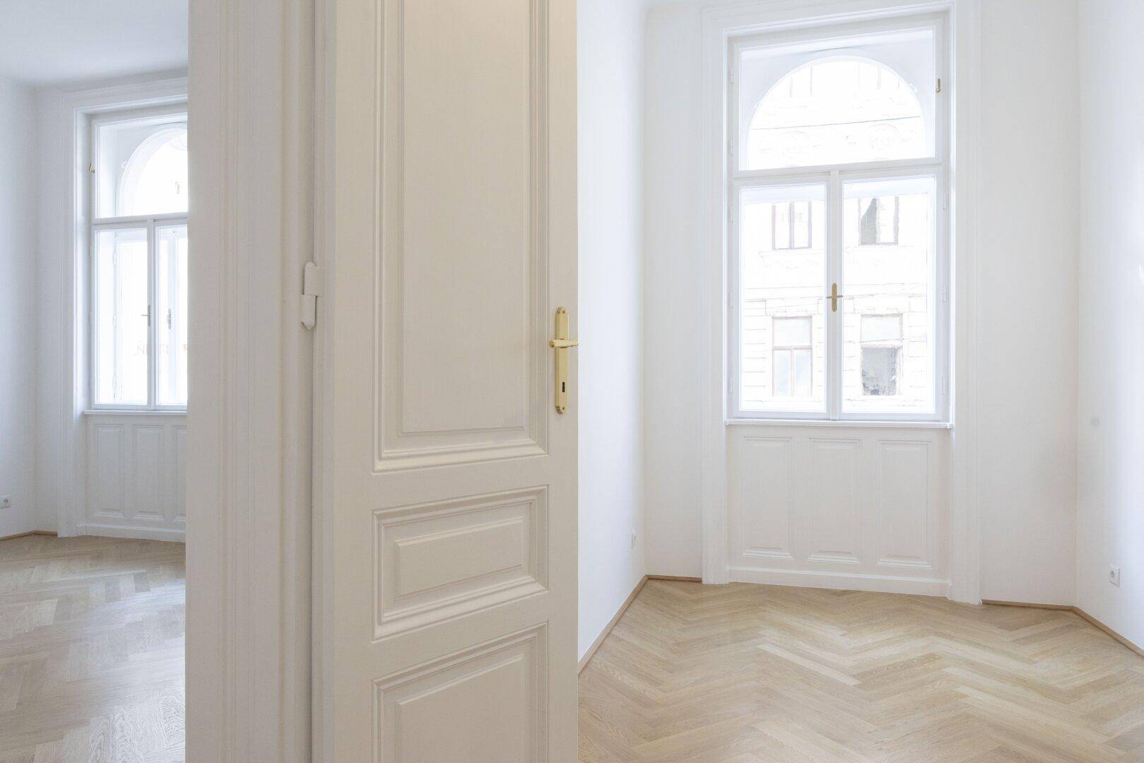 Heller Raum mit Fischgrätparkett und einem großen Bogenfenster, gesehen durch eine offene weiße Tür.