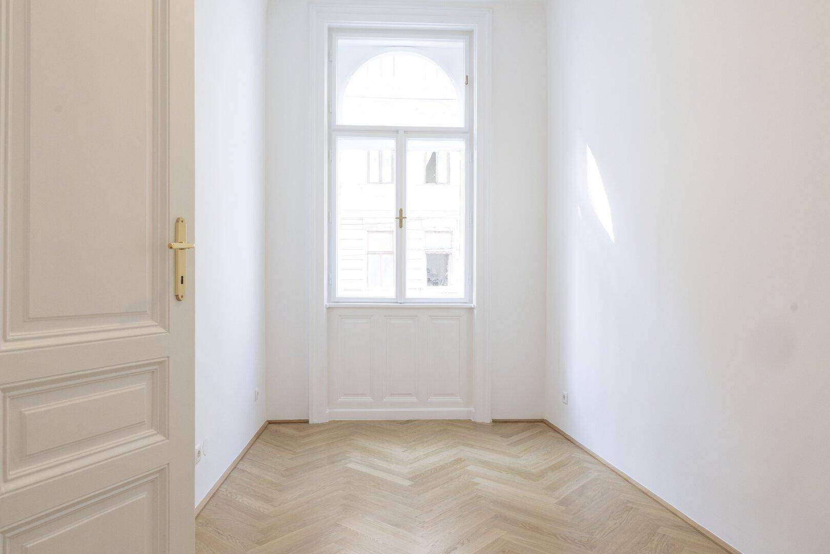 Kleiner, heller Raum mit Fischgrätparkett und einem hohen Bogenfenster mit klassischer Verkleidung.