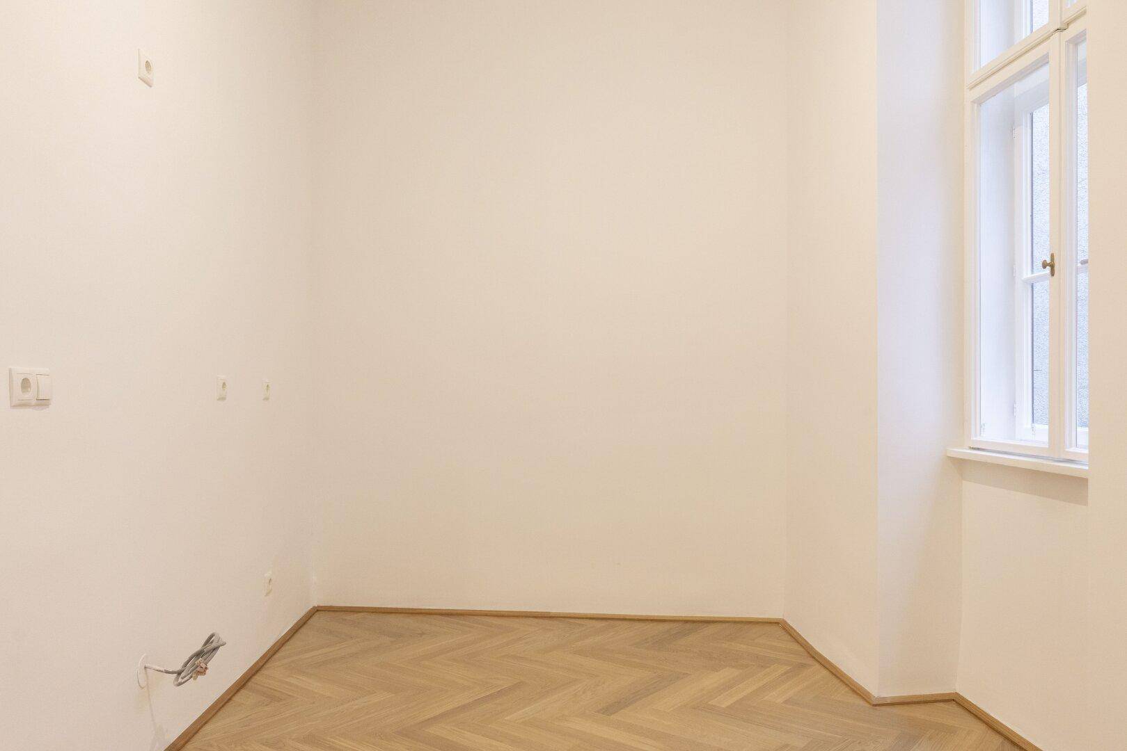 Leerer Raum mit Fischgrätparkett und einem Fenster, das natürliches Licht spendet.