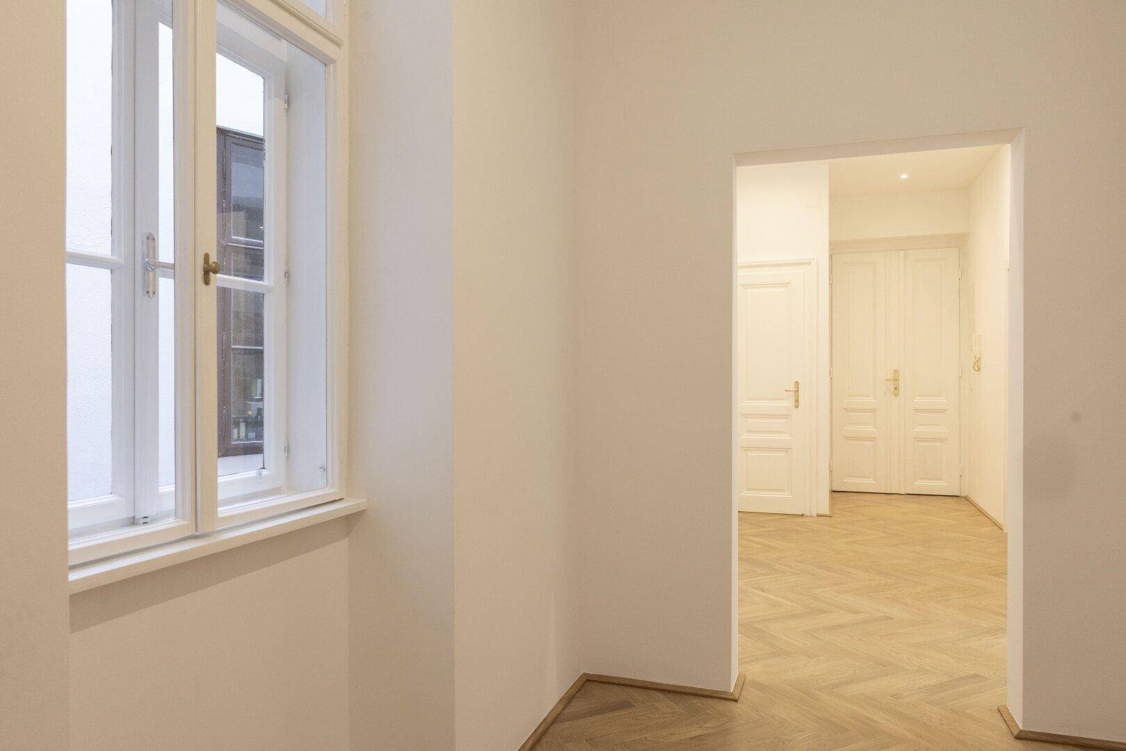 Heller Flur mit Fischgrätparkett und einem Fenster, das Tageslicht hereinlässt.
