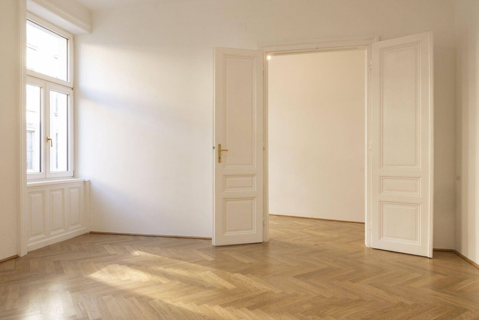 Lichtdurchflutetes Zimmer mit Fischgrätparkett und hohen Doppeltüren, die zu einem weiteren Raum führen.