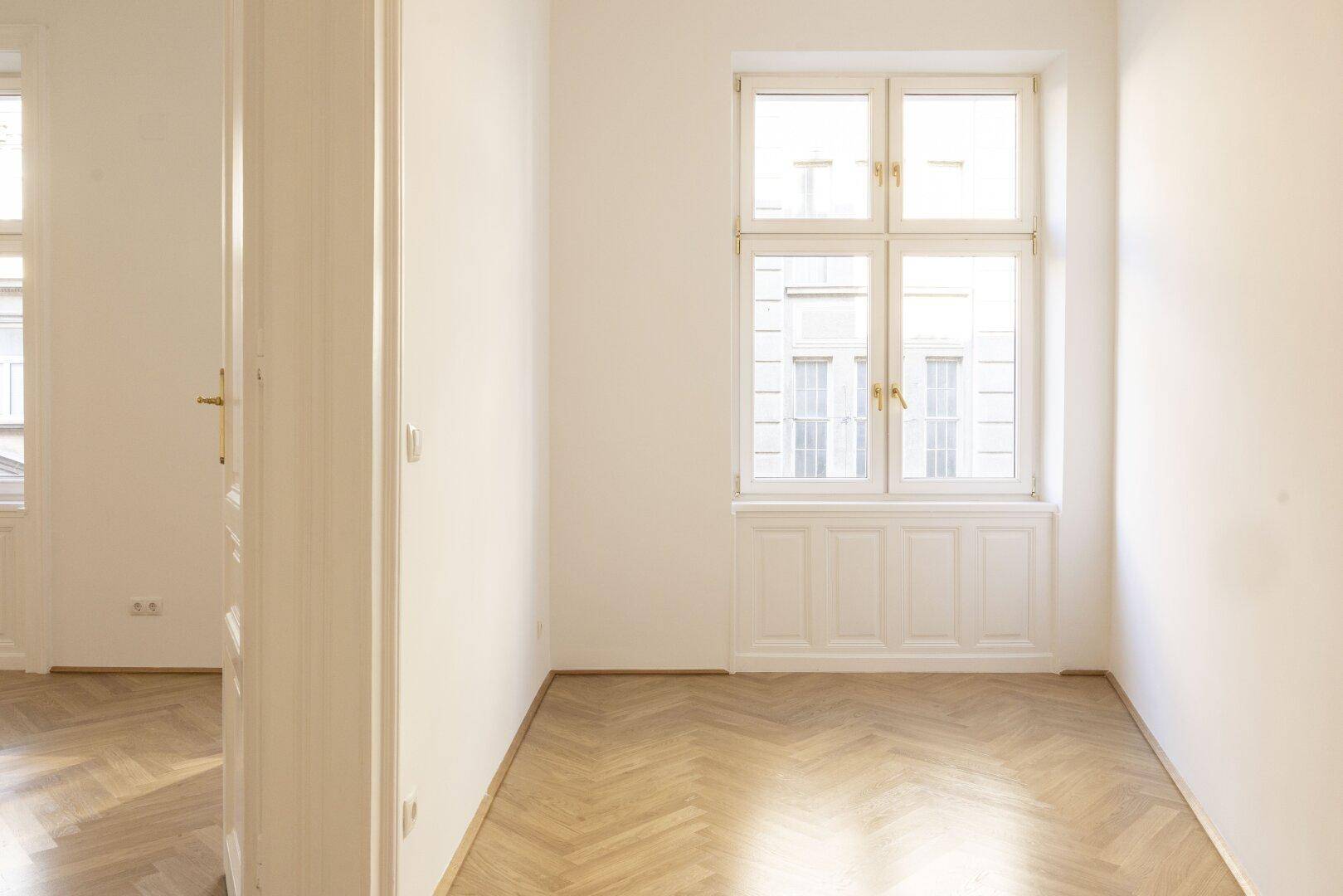 Heller Raum mit Fischgrätparkett und einem großen Fenster, das viel Tageslicht hereinlässt.