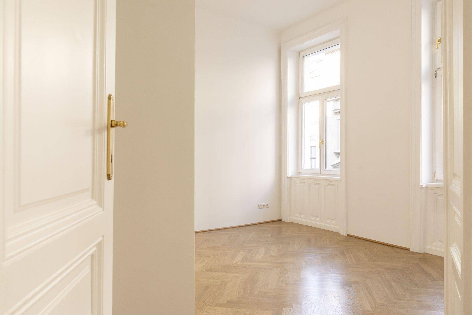 Geräumiges Zimmer mit Fischgrätparkett und hohen Fenstern, das viel natürliches Licht bietet.