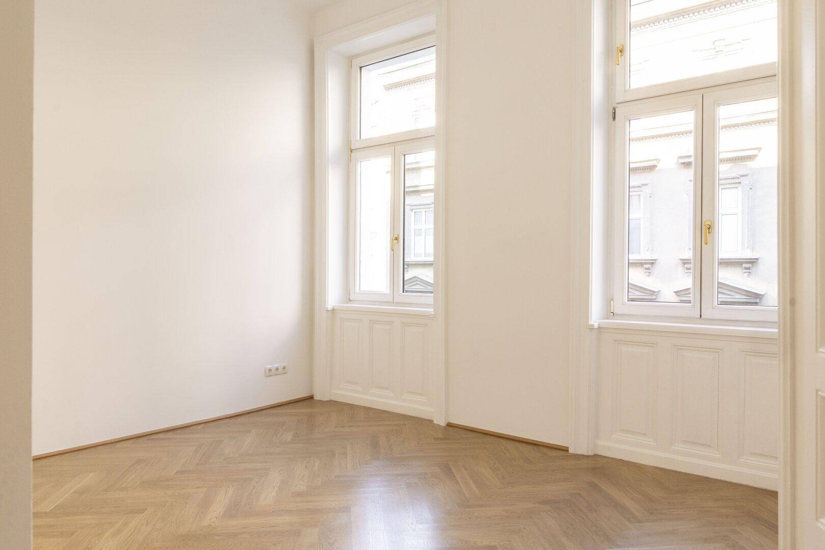 Helles Zimmer mit Fischgrätparkett und zwei hohen Fenstern, die eine angenehme Atmosphäre schaffen.