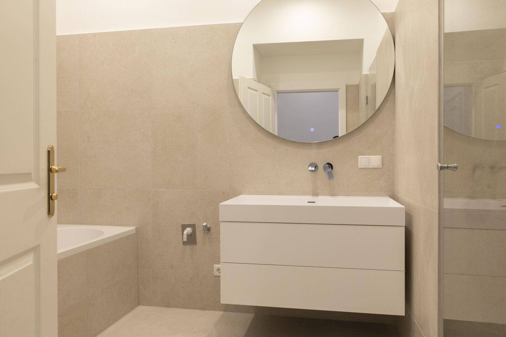 Modernes Badezimmer mit schwebendem Waschtisch, rundem Spiegel und integrierter Badewanne.