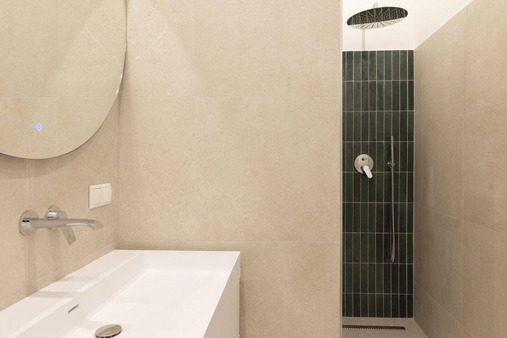 Modernes Badezimmer mit beigefarbenen Fliesen, einer Regendusche und einem runden Spiegel.