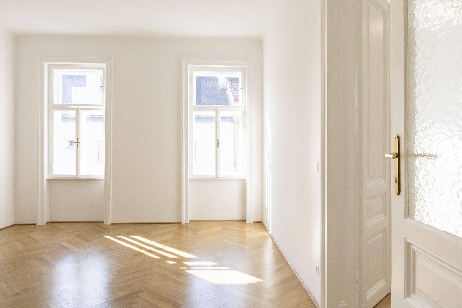 Lichtdurchfluteter Raum mit zwei großen Fenstern, Fischgrätparkett und weißen Wänden, ideal als Wohnzimmer.