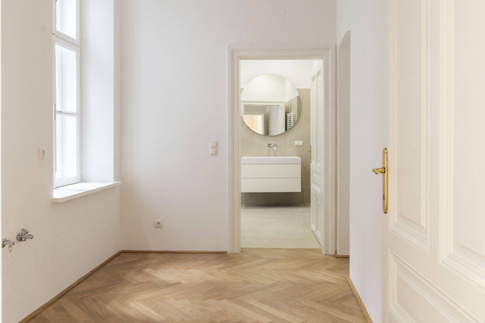 Heller Raum mit Fischgrätparkett und Blick auf ein modernes Badezimmer mit rundem Spiegel und Waschtisch.