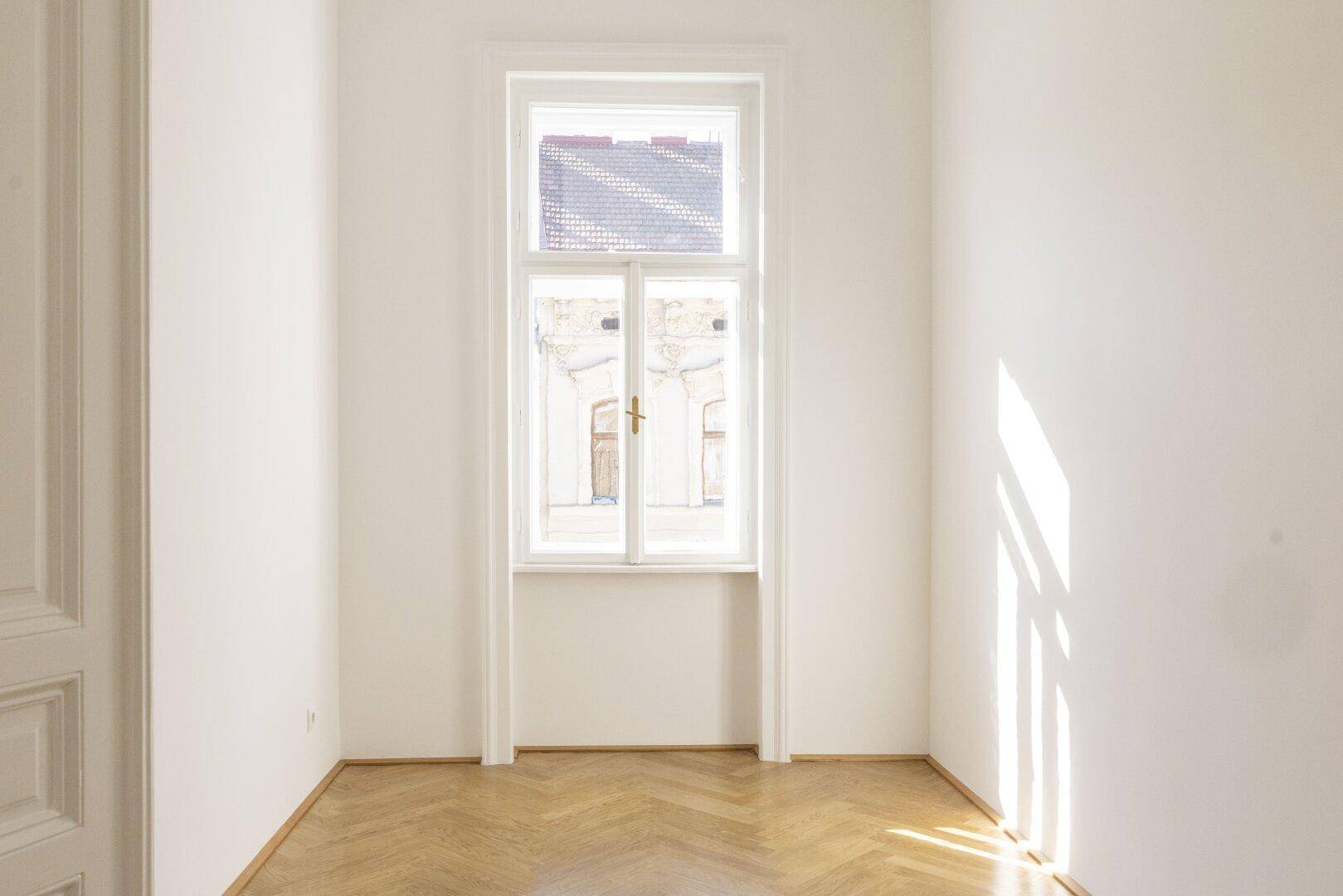 Heller, leerer Raum mit Fischgrätparkett und einem hohen Fenster, das viel Tageslicht hereinlässt.