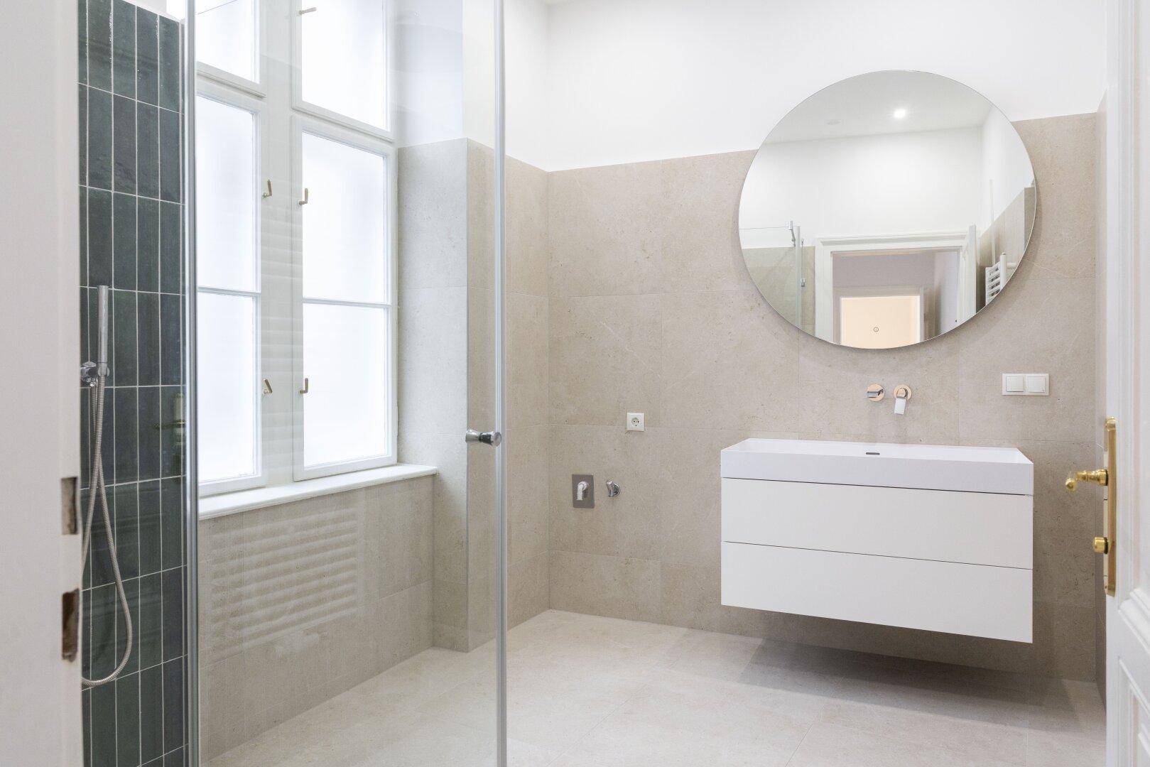 Modernes Badezimmer mit schwebendem Waschtisch, rundem Spiegel, Glasdusche und großem Fenster.