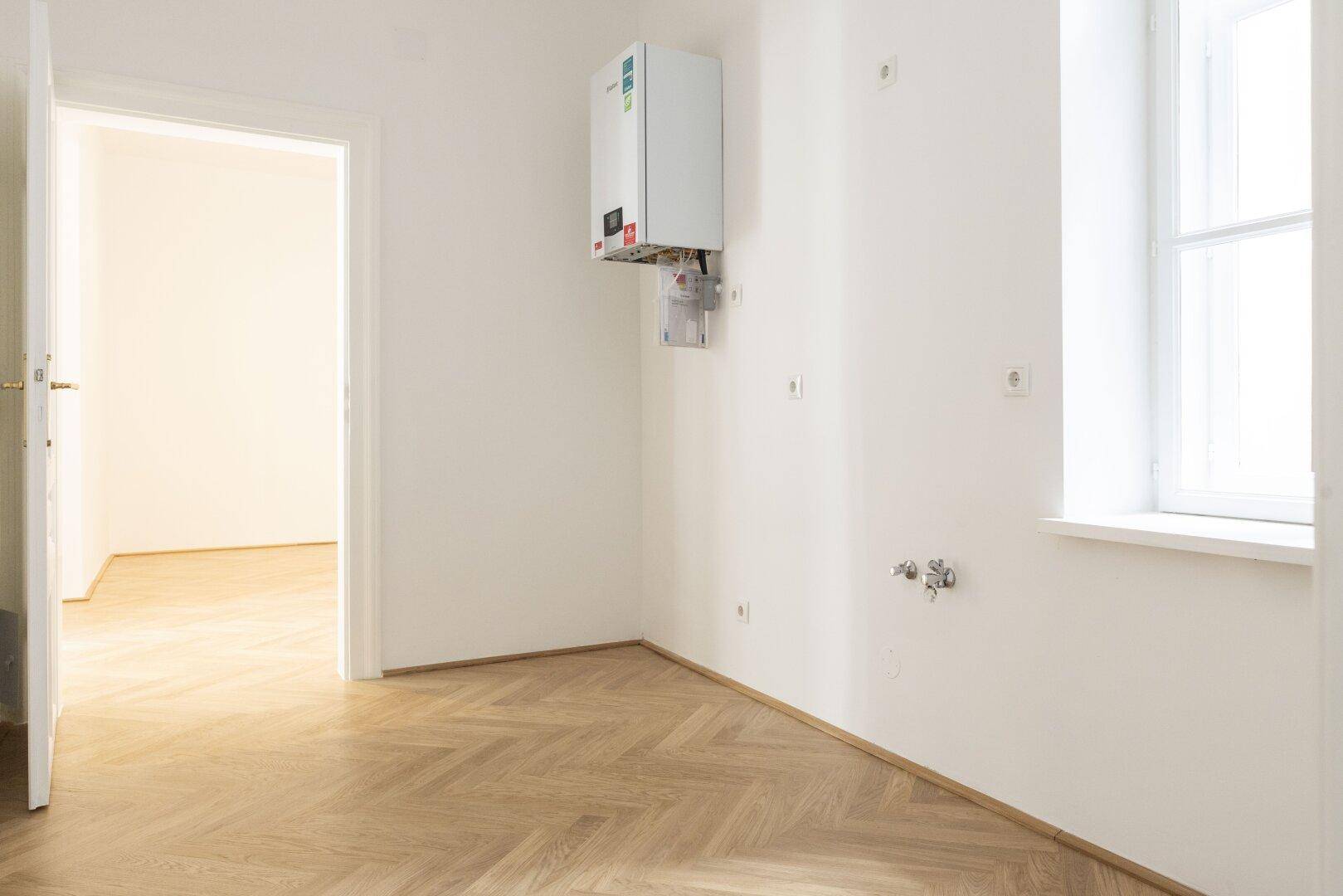Leerer Raum mit Fischgrätparkett, Fenster und einem an der Wand montierten Warmwasserbereiter.