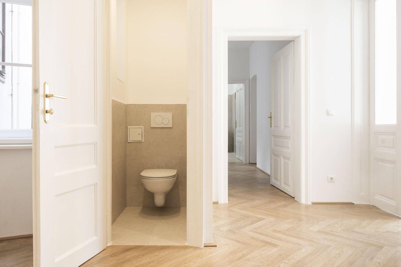 Separates WC mit wandhängender Toilette und modernen Fliesen, angrenzend an einen Raum mit Parkettboden.