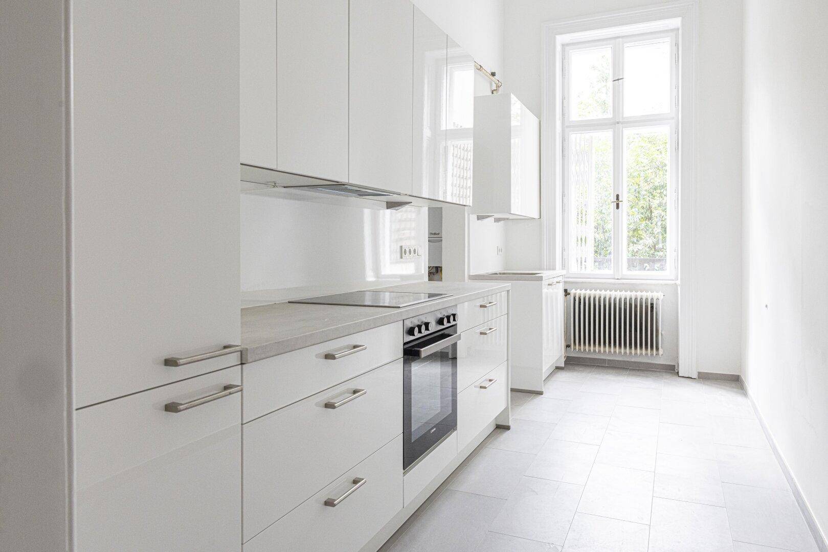 Moderne weiße Küche mit glänzenden Fronten, integrierten Geräten und einem Fenster mit Blick ins Grüne.