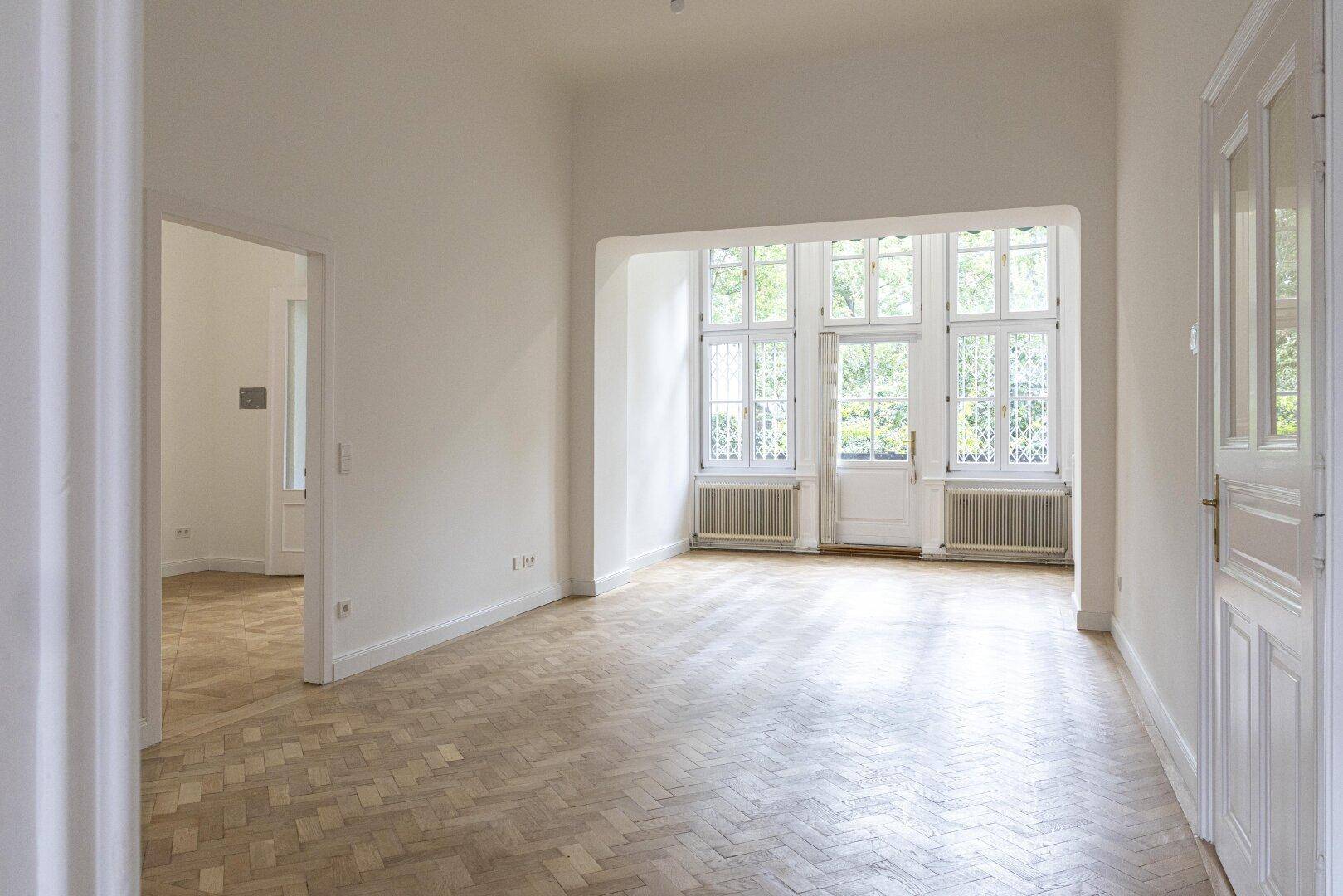 Geräumiger, heller Raum mit Fischgrätparkett und einem großen Erkerfenster, das viel Tageslicht hereinlässt.
