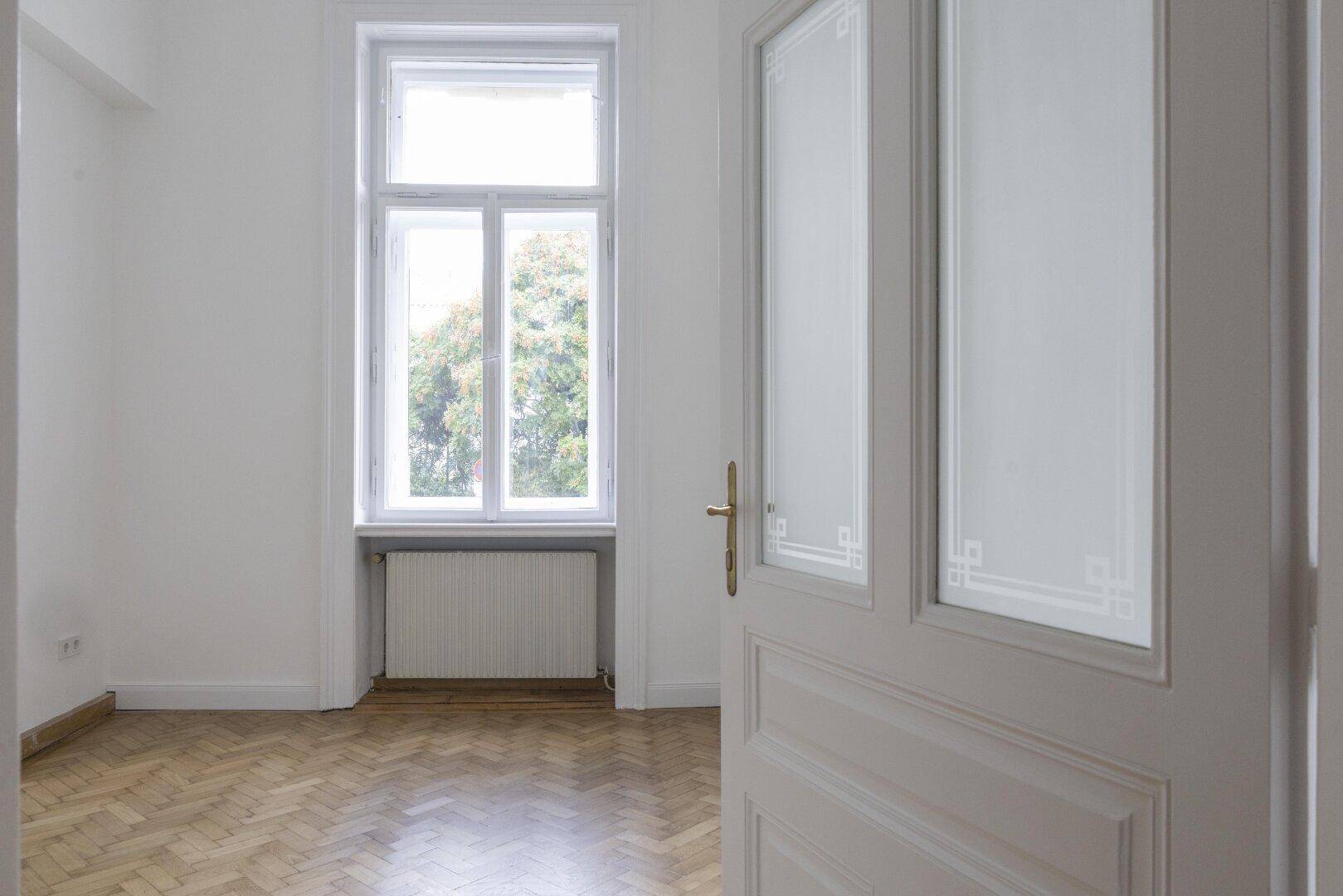 Helles, leeres Zimmer mit Fischgrätparkett und einem großen Fenster, das eine grüne Aussicht bietet.